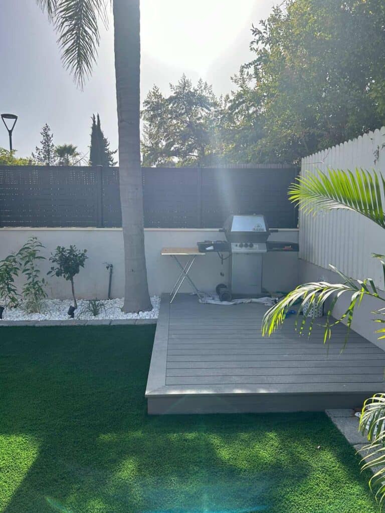 חצר אחורית עם דשא מלאכותי, מרפסת קטנה מוגבהת, גריל גז, שולחן מתקפל וצמחים שונים המוקפים באבנים לבנות וגדר. | DeckTech דקים, פרגולות ועבודות בנייה בעץ