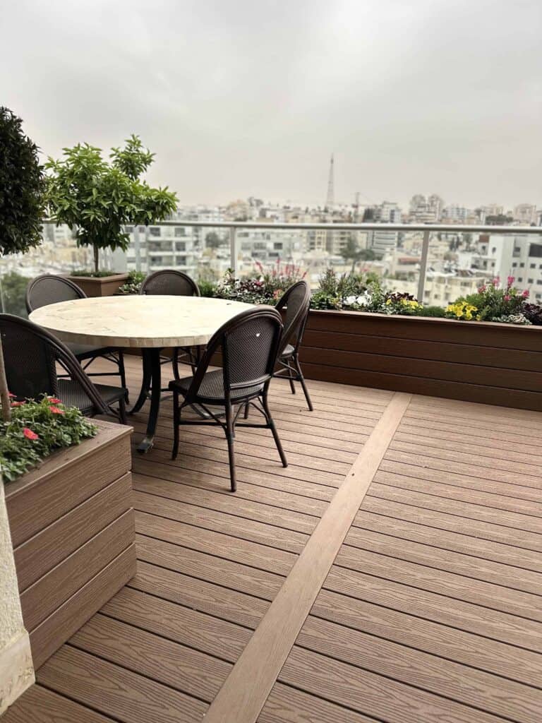 מרפסת גג עם שולחן עגול וארבע כיסאות, מוקפת בעציצים עם פרחים וצמחייה, המשקיפה על נוף עירוני תחת שמים מעוננים. | DeckTech דקים, פרגולות ועבודות בנייה בעץ