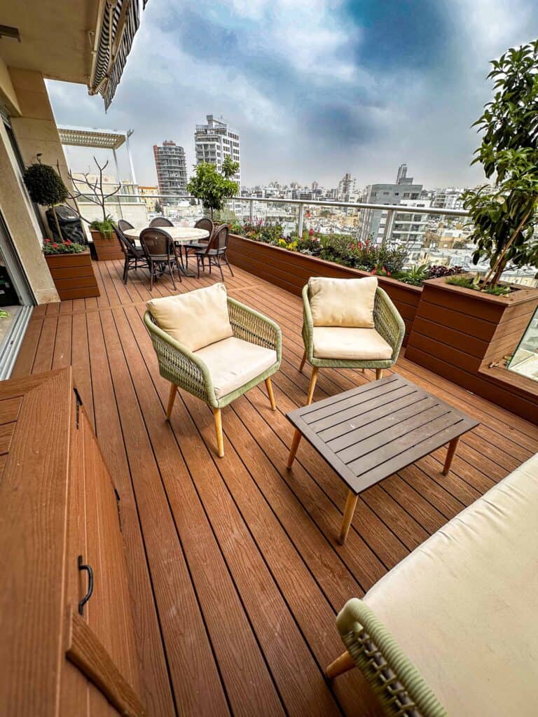 מרפסת גג מודרנית עם ריצוף עץ, כיסאות מרופדים, שולחן, עציצים וקו הרקיע של העיר ברקע תחת שמיים מעוננים. | DeckTech דקים, פרגולות ועבודות בנייה בעץ