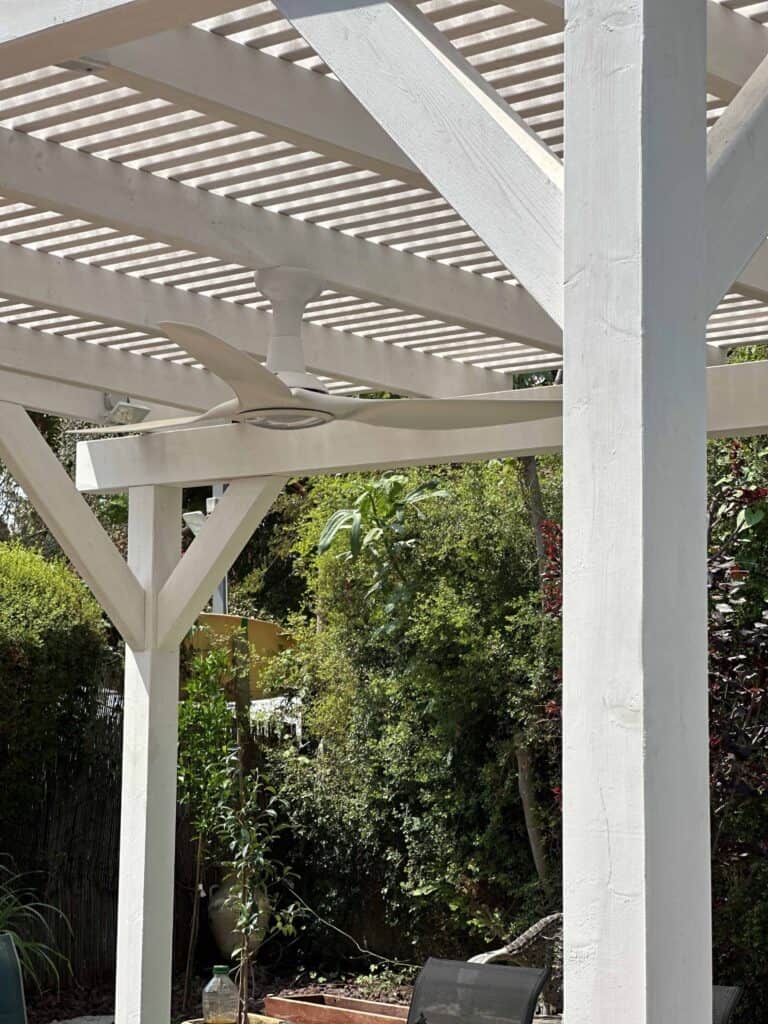 פרגולה מעץ לבן עם מאוורר תקרה מתחתיה, הממוקמת בגינה עם צמחים ירוקים ושיחים ברקע. | DeckTech דקים, פרגולות ועבודות בנייה בעץ