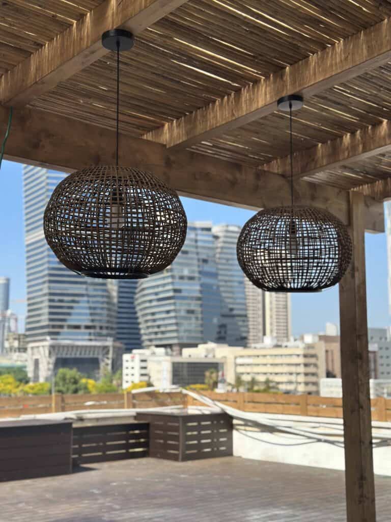 שתי מנורות תלייה כדוריות מקש תלויות מפרגולה מעץ על גג, כשברקע נראים בניינים עירוניים מודרניים. | DeckTech דקים, פרגולות ועבודות בנייה בעץ
