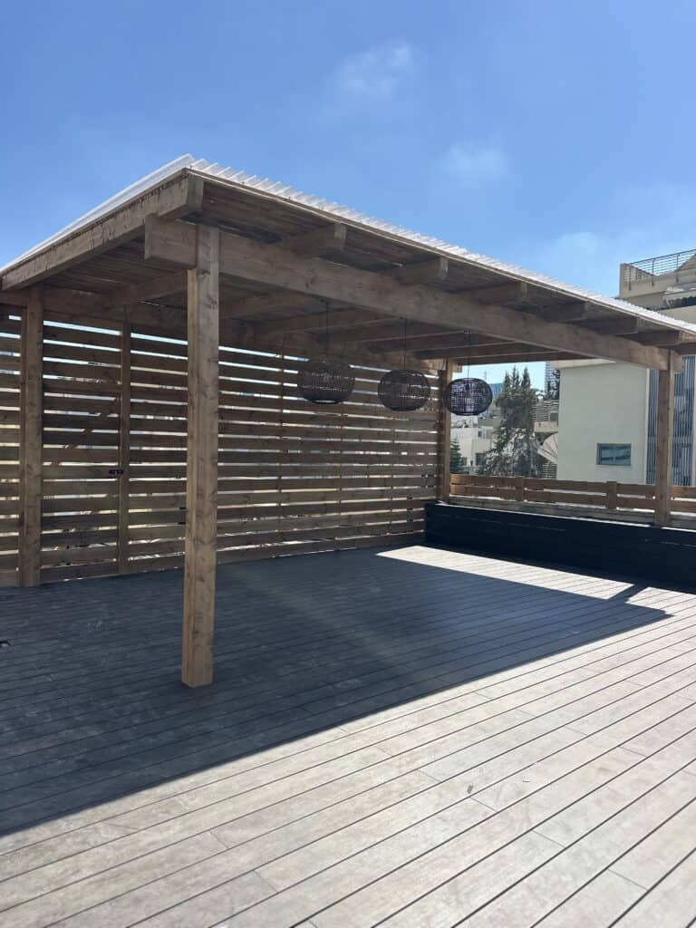פרגולה מעץ עם קירות לוחות ותאורה תלויה על מרפסת מרווחת, תחת שמיים כחולים וצלולים. | DeckTech דקים, פרגולות ועבודות בנייה בעץ
