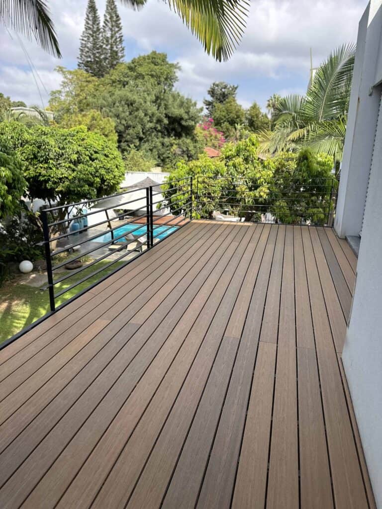 מרפסת עץ עם מעקה מתכת שחור משקיפה על חצר אחורית ירוקה עם עצים, צמחים וחלון ראווה לבריכת שחייה. | DeckTech דקים, פרגולות ועבודות בנייה בעץ
