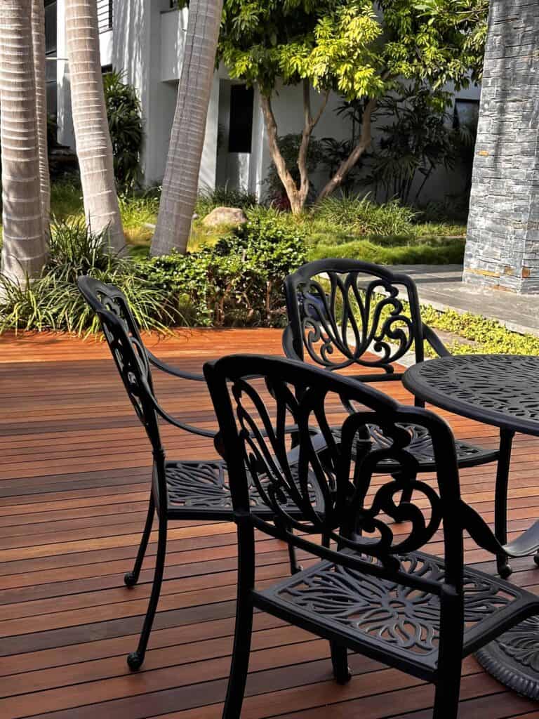 כיסאות מתכת שחורים ושולחן מונחים על דק עץ בחוץ, מוקפים בצמחייה ועצי דקל גבוהים, ובניין נראה ברקע. | DeckTech דקים, פרגולות ועבודות בנייה בעץ