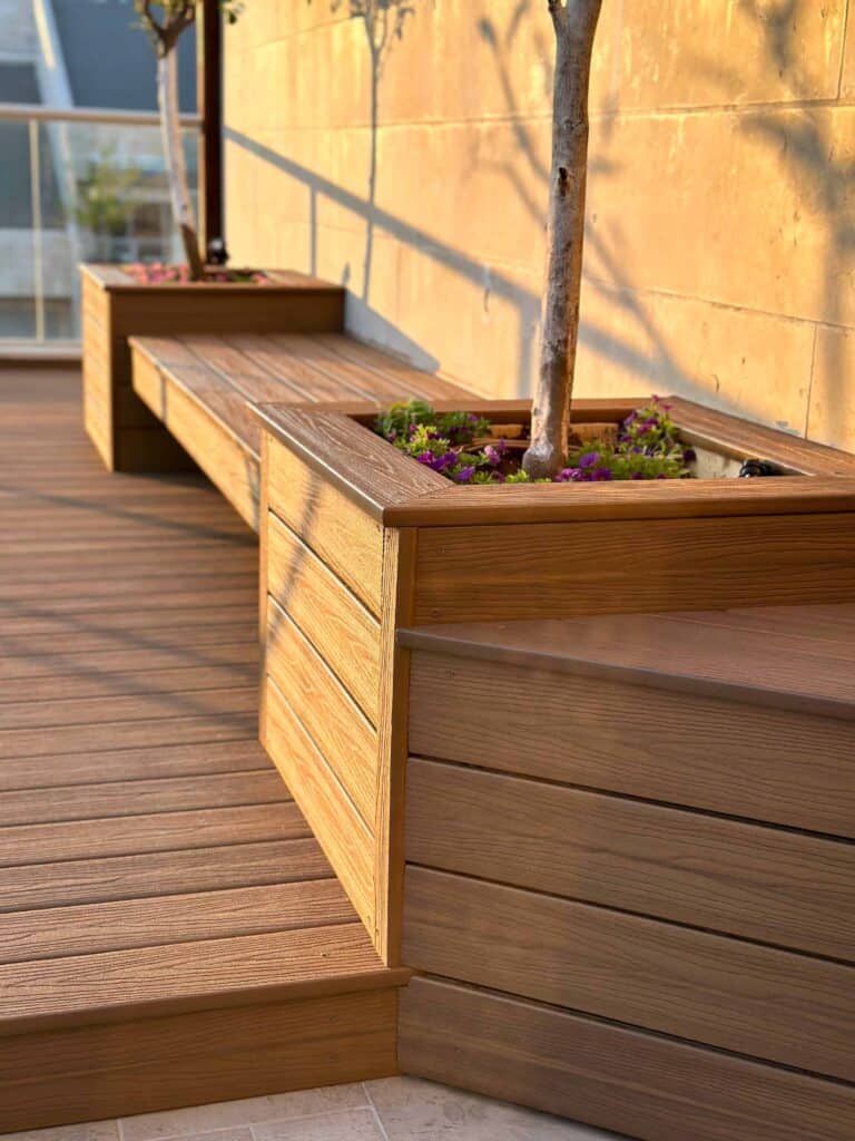 ספסל עץ חיצוני עם אדניות משולבות המכילות עצים קטנים ופרחים, המוצב על דק ליד קיר אבן בצבע בז'. | DeckTech דקים, פרגולות ועבודות בנייה בעץ
