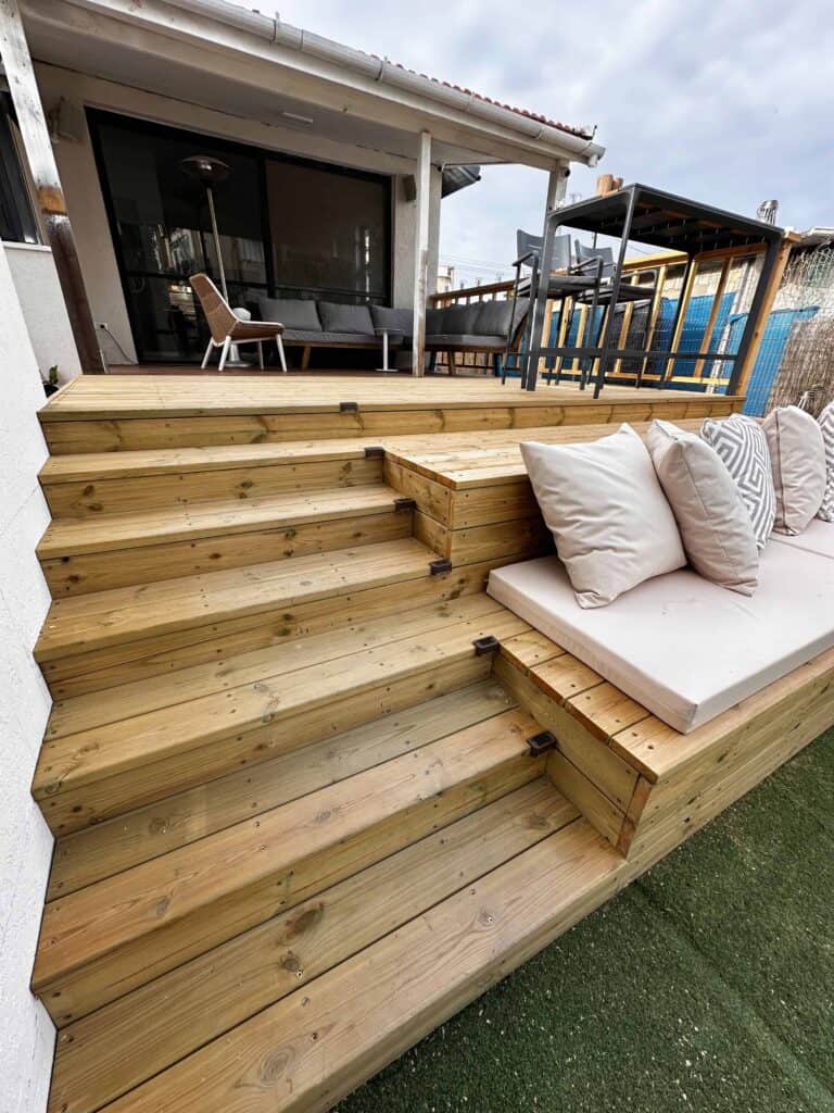 מרפסת עץ עם מדרגות רחבות, ספסל מובנה עם כריות וריהוט גן מול הבית. | DeckTech דקים, פרגולות ועבודות בנייה בעץ