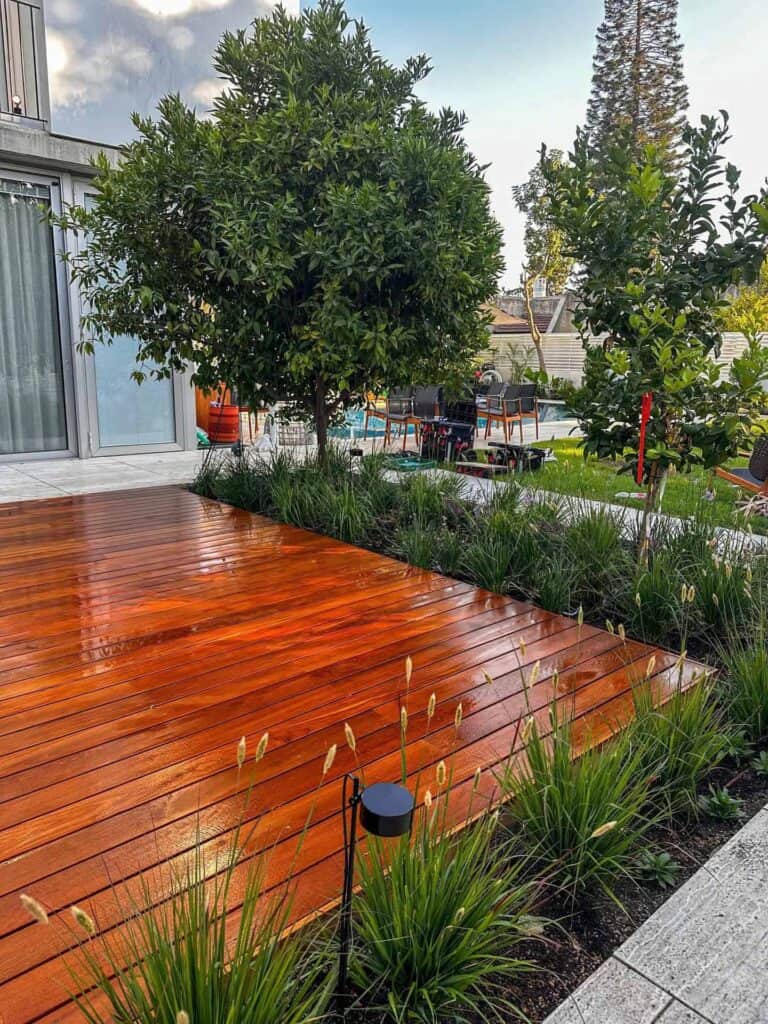 חצר אחורית עם דק עץ, צמחים ירוקים, עצים, ריהוט גן ובריכת שחייה ברקע. | DeckTech דקים, פרגולות ועבודות בנייה בעץ